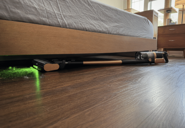 Levoit VortexIQ Pro lying flat under a bed frame, green light revealing cat hair tumbleweeds on hardwood